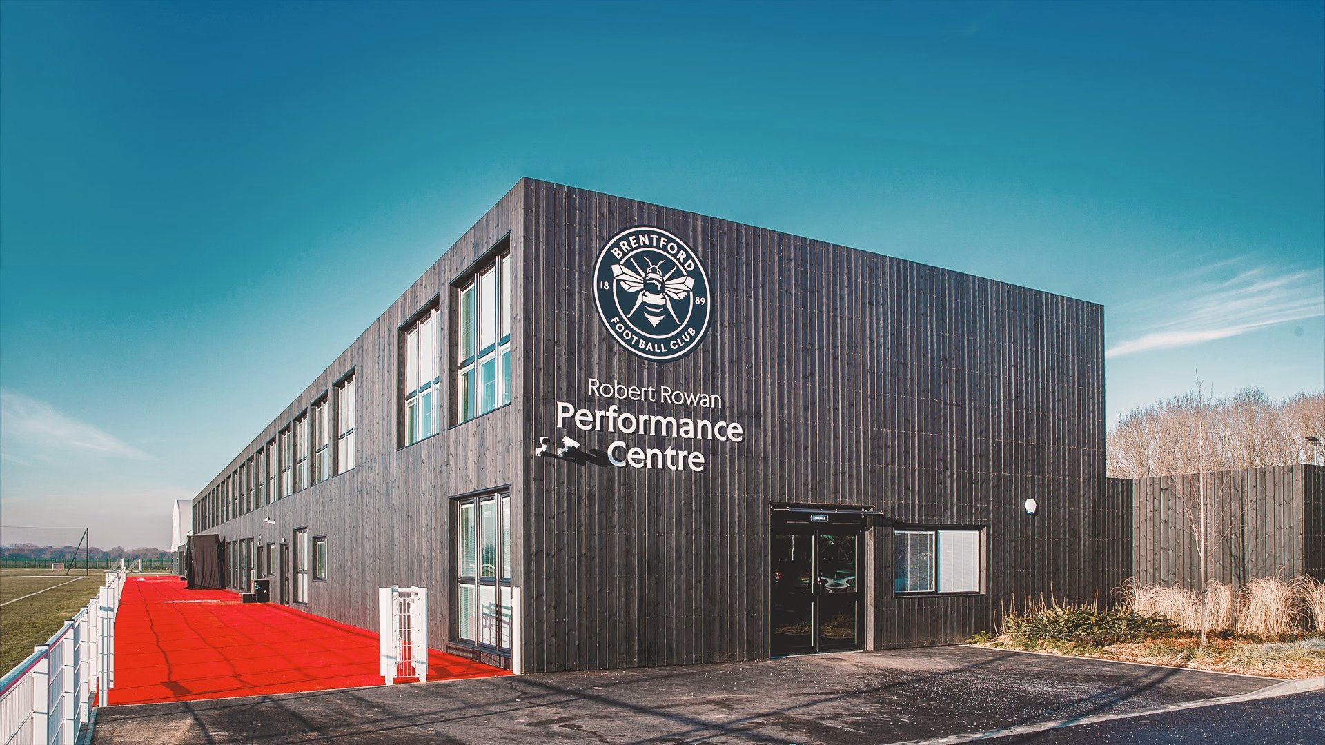 Brentford FC's Robert Rowan Performance Centre, a dark timber-clad building carrying the club crest, beside a training pitch.