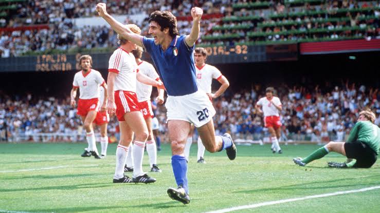 Paolo Rossi in Italy's blue number 20 shirt running with both arms raised in celebration after scoring against Poland at the 1982 World Cup, with Polish players and a packed stadium behind him